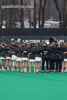 A group of athletes is gathered in a huddle on a sports field, wearing matching uniforms and gear. The ground is green, and there is signage with a brand logo in the background. Trees and parked cars can be seen behind a fence surrounding the field. A person on the left side is capturing the moment with a camera.
