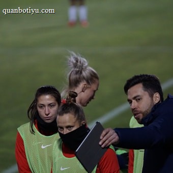 A group of individuals on a sports field are wearing green vests over red attire. One person is holding a tablet and seems to be discussing or showing something to the others. The atmosphere suggests a sports event, likely soccer, indicated by their clothing and the setting. The background is a grassy field.