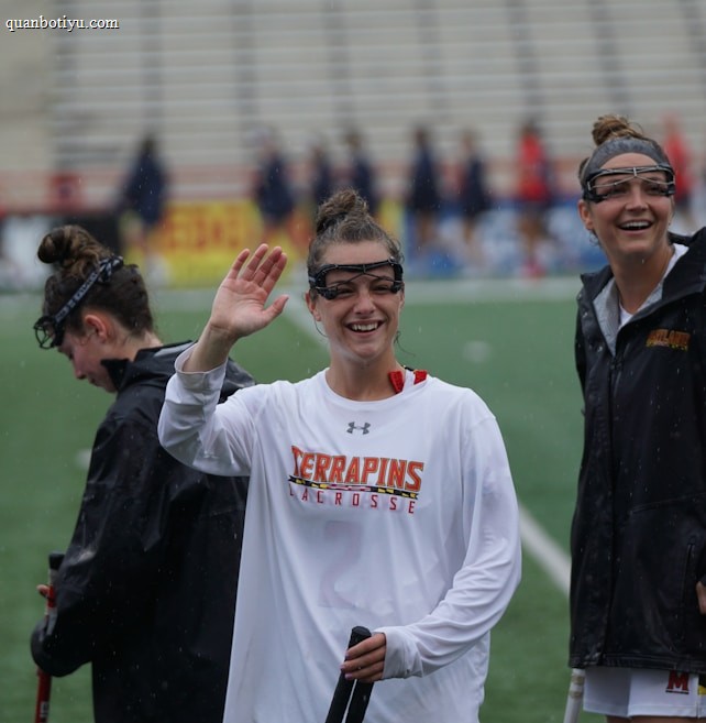 Several athletes in sports attire are on an outdoor sports field, interacting and smiling. One individual in the foreground waves with a friendly expression, while another stands beside her, also smiling. They wear protective goggles and gear associated with lacrosse. The background features indistinct figures in dark clothing against a backdrop of stadium seating.